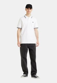 Polo bianco con accenti a righe nere e bianche, abbinato a jeans scuri e scarpe da ginnastica nere. Il modello indossa un cappellino nero.