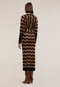 Langes Kleid mit Chevron-Muster in Braun und Schwarz, das lange Ärmel und eine taillierte Passform hat, kombiniert mit glänzenden braunen Stiefeln mit Absatz.
