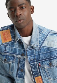 Jeansjack met een patchworkontwerp in lichte en donkere blauwtinten, koperen hardware en een leren label met merknaam op de linkerschouder.