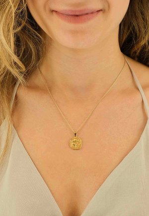 Collier pendentif en or avec un charme texturé de forme irrégulière sur une chaîne délicate, porté sur un haut de couleur claire.