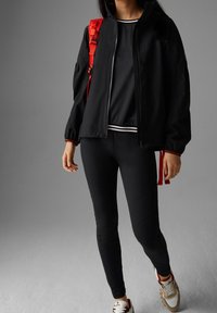 Černá větrovka s červeným zipem a detaily, v kombinaci s černými legínami s texturovanými panely a teniskami s vícebarevnými detaily.