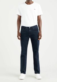 Mörkblå jeans med klassisk passform, kombinerade med en vit t-shirt med rund hals och en liten röd logga på bröstet. Bärs med vita sneakers.