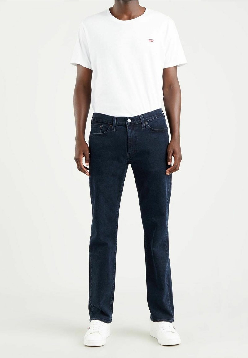 Mörkblå jeans med klassisk passform, kombinerade med en vit t-shirt med rund hals och en liten röd logga på bröstet. Bärs med vita sneakers.