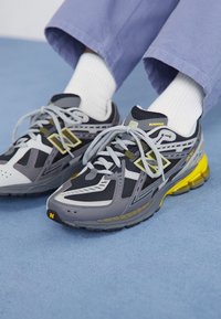 Chaussures de course grises avec des accents noirs et jaunes. Tige en mesh et synthétique, semelle épaisse, design à lacets et panneaux texturés distincts.