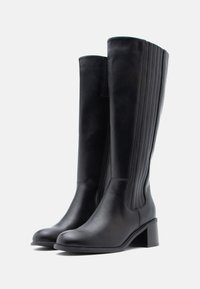 Fekete bőrből készült, térd felett érő csizmák sima felülettel, függőleges bordázattal az oldalán és tömbsarkú kialakítással.