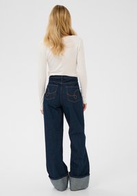 Mørkeblå jeans med høj talje og vidde ben, der har opkrogede manchetter og dekorative syninger på baglommerne.