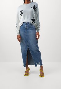 Weißer gestrickter Pullover mit schwarzem Blumenapplikation, kombiniert mit einem blauen Denim-Midirock mit einem Schlitz vorne und fransigem Saum. Gelbe Riemchen-Sandalen.