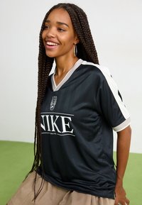 Svart Nike sportsjersey med korte ermer, grå og hvite detaljer, og v-hals. Har en logo og horisontal tekst over foran.