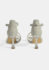 Zilveren glitter hoge hakken sandalen met enkelbandjes, open achterkant, gebogen hak en subtiele decoratieve kraaldetails langs de randen.