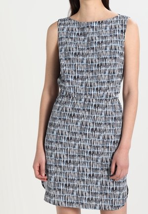 Nainen yllään hihaton polvipituinen mekko, jossa on sinisen, mustan ja valkoisen abstrakti pystysuora raidallinen kuvio, seisomassa yksinkertaista vaaleaa taustaa vasten.