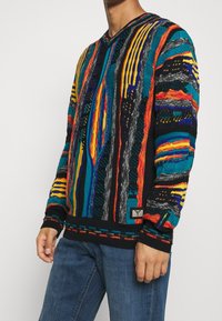 Värikäs neulepaita, jossa on V-kaula-aukko, erilaisia kuvioita punaisessa, oranssissa, sinisessä ja mustassa, tekstuuriset korostukset ja etiketti helman kohdalla.