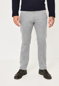 Light grey cotton trousers with a straight leg design, paired with black lace-up shoes. Model stands with hands by sides, wearing a navy sweater.