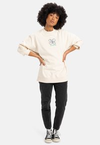 Beżowy, oversize'owy sweatshirt z okrągłą grafiką w zieleni i niebieskim, w połączeniu z czarnymi spodniami typu tapered i czarnymi trampkami z białą podeszwą.