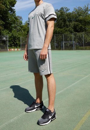 Homme en t-shirt et shorts Nike gris avec des baskets Nike noir et blanc se tient sur un terrain de sport extérieur sous le soleil.