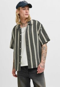 Camisa de manga corta, con botones, en un patrón de rayas en blanco y negro, que presenta un corte relajado y una tela ligera. Llevada sobre una camiseta blanca de canalé.