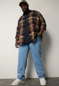 Camicia di flanella a quadri nei toni del verde, rosso e marrone, indossata sopra una t-shirt nera. Jeans azzurri chiari e sneakers bianche completano il look.