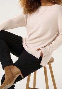 Maglione a coste beige chiaro con scollo ampio, abbinato a pantaloni neri aderenti e stivali alla caviglia in pelle scamosciata marrone, su uno sgabello di legno.