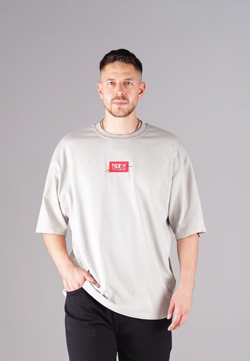 Light gray oversized t-shirt, short sleeves, round neckline, featuring a red brand logo patch on the front. Worn with dark pants.