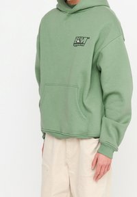 Sudadera con capucha verde y un bolsillo frontal, con un logo gráfico negro en el pecho. La tela parece suave y texturizada.