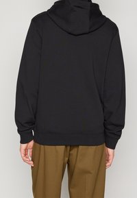 Felpa nera con cappuccio e tasca a marsupio, con polsini a coste. Tessuto morbido con una vestibilità comoda, abbinata a pantaloni larghi di colore marrone chiaro.