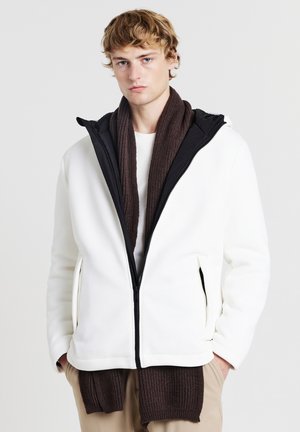 Giacca bianca con zip a coste e cappuccio nero, abbinata a una sciarpa marrone a coste. Presenta tasche laterali e una vestibilità rilassata.