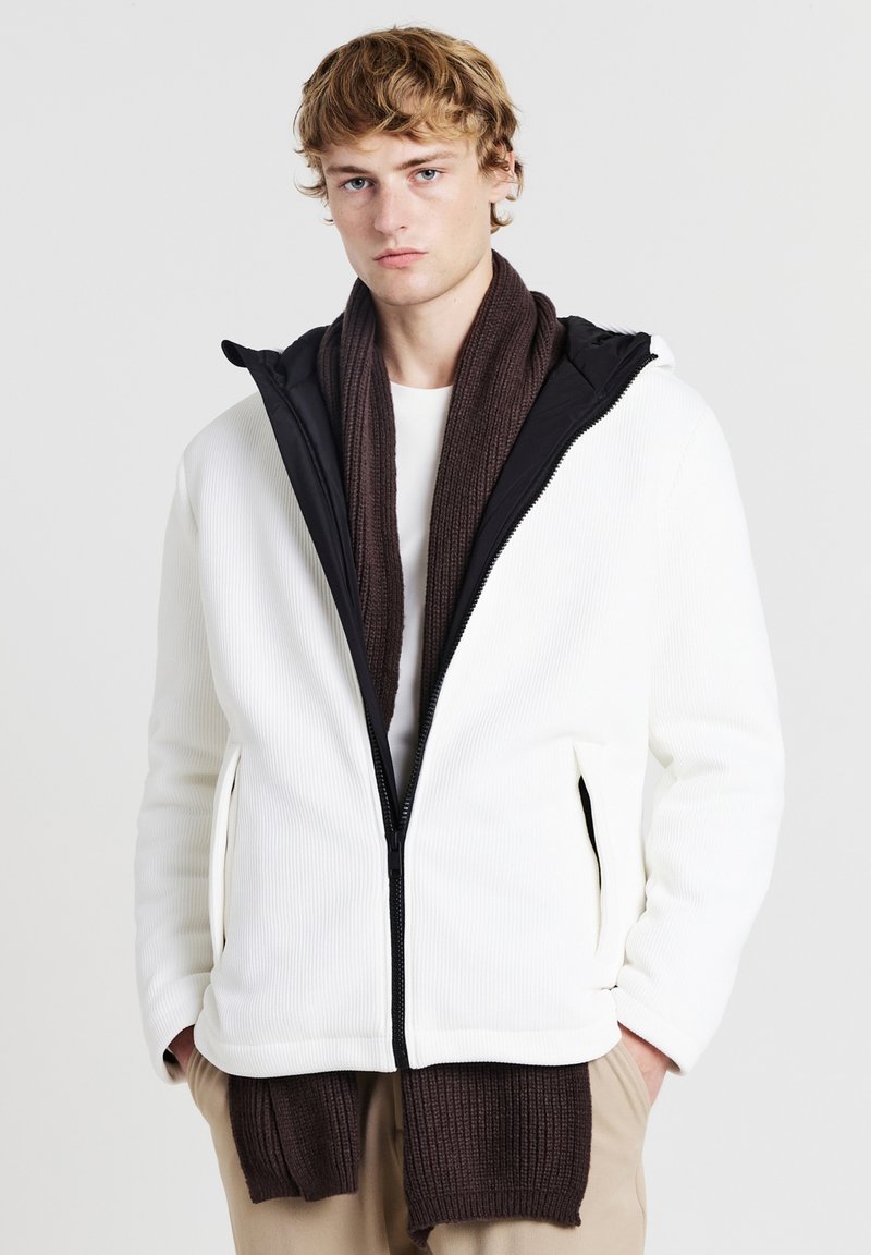Weißer, gerippter Reißverschluss-Jacke mit schwarzer Kapuze, kombiniert mit einem braunen, gerippten Schal. Verfügt über seitliche Taschen und eine lockere Passform.