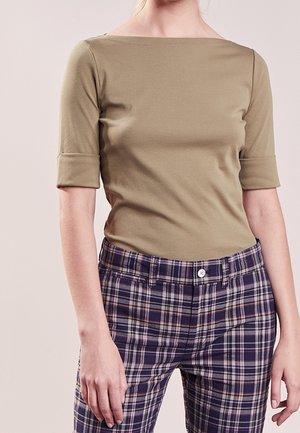 Kobieta w oliwkowej bluzce z rękawem do łokci i spódnicach w krótkim kroju w niebieską kratę, stojąca na jednolitym tle.