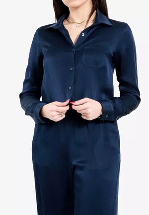 Camicetta in satin blu navy con maniche lunghe, chiusura a bottoni sul davanti, taschino singolo sul petto e una consistenza liscia e lucida.