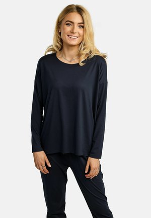 Blondínka sa usmieva, má na sebe voľnú čiernu blúzu s dlhým rukávom a čierne nohavice, stojí pred jednoduchým bielym pozadím.