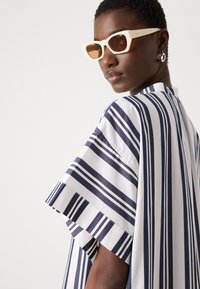 Camicia a righe bianca e blu navy, vestibilità rilassata con maniche larghe, dotata di scollo rotondo e tessuto testurizzato. Gli accessori includono occhiali da sole angolari.