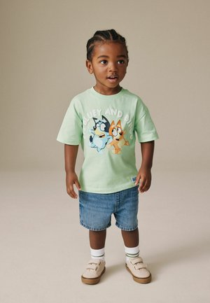 Ungt barn med flettet hår iført en lys grøn Bluey og Bingo t-shirt, denimbukser, hvide sokker og beige sneakers, stående.