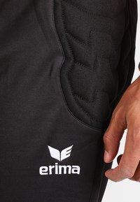 Schwarze Sporthosen mit strukturiertem Polster an der Seite. Weißes "erima"-Logo am unteren Ende. Slim Fit mit glattem Stoff.