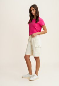 T-shirt rose avec impression graphique, shorts en coton crème avec poches latérales, et baskets blanches avec semelles marron clair. Design simple et décontracté.