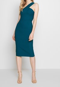 Robe midi ajustée teal avec une bretelle, détails plissés et jupe droite. Fabriquée en tissu extensible, accompagnée de talons nus à bride autour de la cheville.