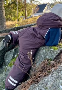 Lilla vandtætte bukser med hvide striber og en blå jakke, iført mens jeg læner mig op ad en sten i et skovområde med fyrnåle og mos.