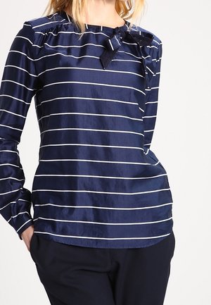 Vrouw draagt een marineblauwe blouse met lange mouwen en witte horizontale strepen, met een gestrikte strik bij de hals, gecombineerd met een zwarte broek.