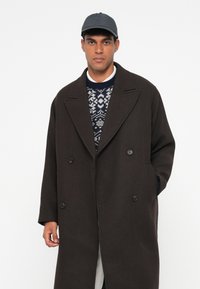 Uomo che indossa un cappotto doppiopetto marrone scuro, un maglione blu con motivo, una camicia bianca, pantaloni chiari e un cappellino da baseball grigio scuro, in piedi di fronte a uno sfondo bianco.