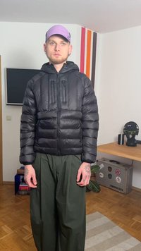 Čierna nadýchaná bunda s kapucňou, vybavená dvoma náprsními vreckami a zapínaním na zips. Kombinovaná s voľne strihanými olivovými zelenými nohavicami.