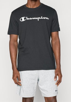 Homme portant un t-shirt noir Champion avec un logo blanc et un short Champion gris clair sur un fond uni.