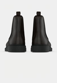 Pár tmavě hnědých kožených Chelsea bot zobrazených zezadu, vybavených černými elastickými bočními panely a silnými černými podrážkami na bílém pozadí.
