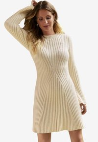 Beige neulemekko, jossa on pystysuora ribbaus, pyöreä pääntie ja pitkät hihat. Tiukka yläosa levenee helmaan. Teksturoitu kankaan yksityiskohta.