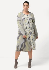 Stickad cardigan i grått med olivgrön och lavendelfärgad vågmönster, lagd över en plisserad klänning med abstrakt gult och svart mönster.