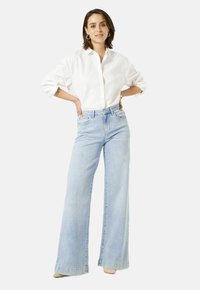 Vrouw met kort bruin haar, gekleed in een witte blouse met lange mouwen, lichtblauwe wijde jeans, beige schoenen en gouden creolen, die met haar handen op de heupen staat.