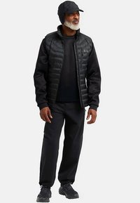 Schwarze isolierte Weste über einem schwarzen Langarmshirt, kombiniert mit schwarzer Hose und passender Mütze. Strukturierter Stoff, praktisches Design.