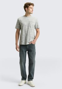 Grijs gestreept T-shirt met korte mouwen en een borstzak, gecombineerd met blauwe jeans en witte sneakers, staand tegen een lichte achtergrond.