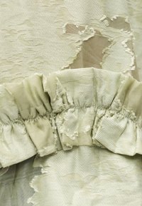 Κοντινό πλάνο ανοιχτόχρωμου πράσινου υφάσματος με υφασμένα, ξεφτισμένα ανθεκτικά μοτίβα λουλουδιών και λεπτομέρεια μαζεμένης βολάν περιθωρίου.