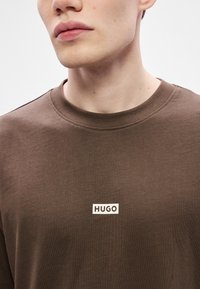 Brązowy bawełniany t-shirt z okrągłym dekoltem i białym prostokątnym logo z napisem "HUGO" na piersi. Gładka tekstura, minimalistyczny design.