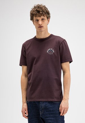 Man met krullend haar die een donkerpaarse korte mouw T-shirt draagt met een klein wit logo op de linkerborst en blauwe spijkerbroek.