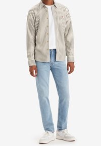 Camisa de botones a rayas en beige y verde oliva, combinada con jeans de color azul claro y zapatillas blancas. Presenta un logo en el pecho y botones dorados.