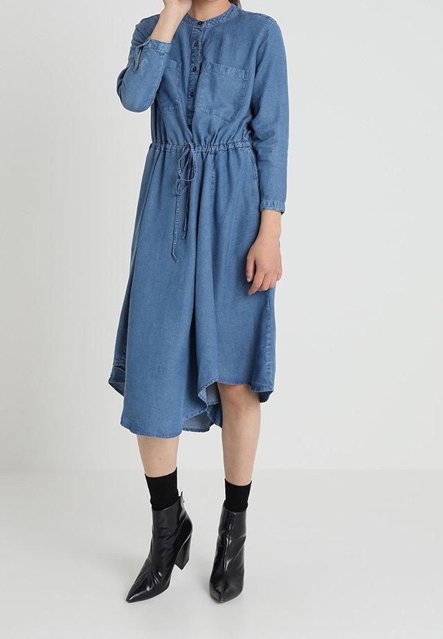 Robe en denim à manches longues, à boutons sur le devant, avec une taille ajustable par cordon. Elle possède une poche sur le côté gauche et un ourlet arrondi. Associée à des bottines noires.